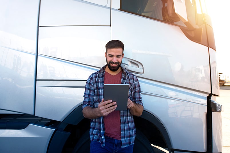 A semi-truck driver leans against his vehicle reviewing telematics data on tablet.